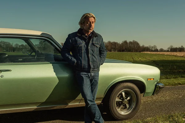 stock image Man with blond hair in jeans stands on front side of a vintage american muscle car in sunny countryside.