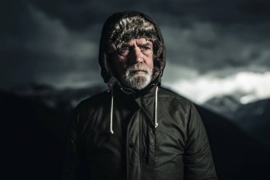 Gri sakallı yeşil ceketli kıdemli dağcı karlı dağ manzarasında duruyor..