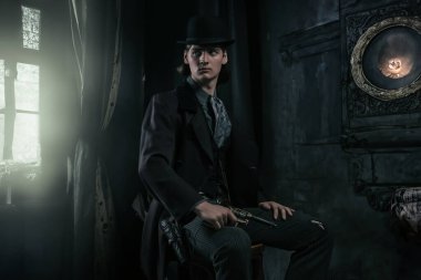 Şapkası ve paltosu olan genç Viktorya 'lı bir adam bir odada elinde tabanca olan bir sandalyede oturuyor..