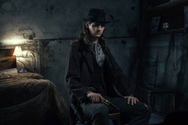 Şapkası ve paltosu olan genç Viktorya 'lı bir adam bir odada elinde tabanca olan bir sandalyede oturuyor..