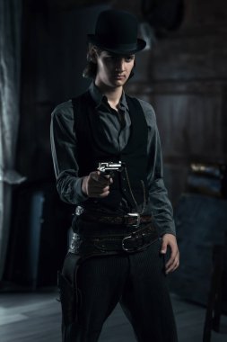 Şapkalı genç bir Victoria 'lı bir adam bir odada durmuş tabancasını doğrultuyor..