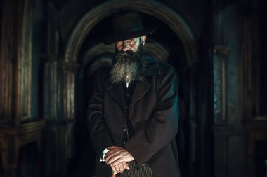 Şapkalı ve paltolu muhafazakar adam eski koridorda bastonuyla dikiliyor..