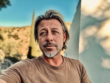 Kahverengi tişörtlü mutlu sarışın adam güneşli kırsalda beyaz bir evin yanında.