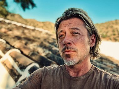 Kahverengi tişörtlü mutlu sarışın adam güneşli kırsalda beyaz bir evin yanında.