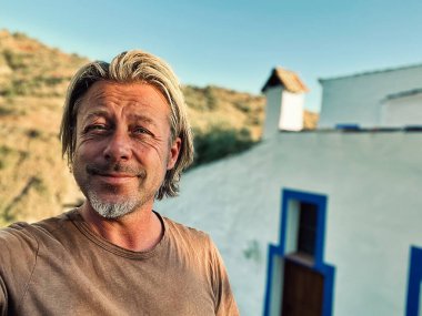 Kahverengi tişörtlü mutlu sarışın adam güneşli kırsalda beyaz bir evin yanında.