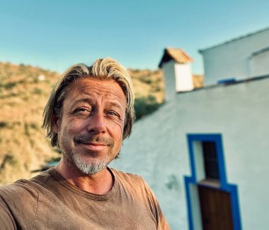 Kahverengi tişörtlü mutlu sarışın adam güneşli kırsalda beyaz bir evin yanında.