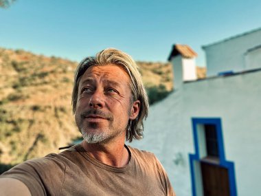 Kahverengi tişörtlü mutlu sarışın adam güneşli kırsalda beyaz bir evin yanında.