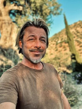 Kırsal kesimde, yaşlı bir zeytin ağacının yanındaki kahverengi tişörtlü mutlu sarışın adam..