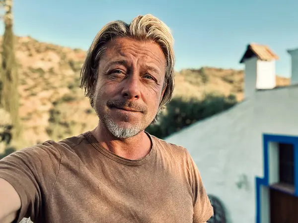 Kahverengi tişörtlü mutlu sarışın adam güneşli kırsalda beyaz bir evin yanında.