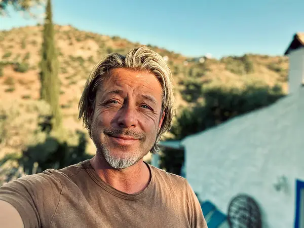 Kahverengi tişörtlü mutlu sarışın adam güneşli kırsalda beyaz bir evin yanında.