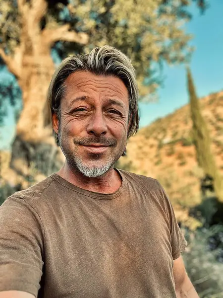 Kırsal kesimde, yaşlı bir zeytin ağacının yanındaki kahverengi tişörtlü mutlu sarışın adam..