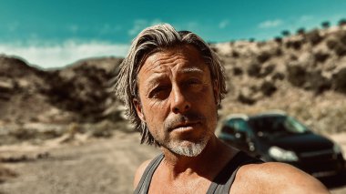 Bronz tenli, sakallı, atlet gömlekli, güneşli bir ortamda, arka planda siyah bir araba olan sarışın bir adam..