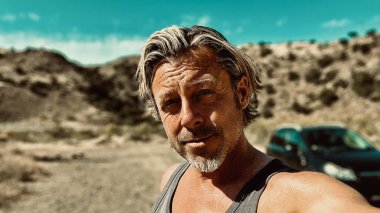 Bronz tenli, sakallı, atlet gömlekli, güneşli bir ortamda, arka planda siyah bir araba olan sarışın bir adam..