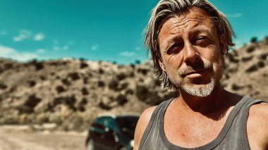 Bronz tenli, sakallı, atlet gömlekli, güneşli bir ortamda, arka planda siyah bir araba olan sarışın bir adam..