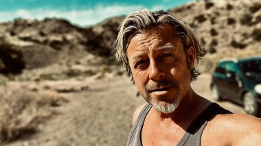 Bronz tenli, sakallı, atlet gömlekli, güneşli bir ortamda, arka planda siyah bir araba olan sarışın bir adam..