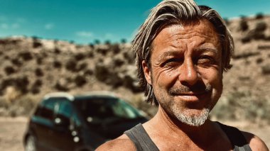 Bronz tenli, sakallı, atlet gömlekli, güneşli bir ortamda, arka planda siyah bir araba olan sarışın bir adam..