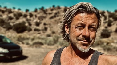 Bronz tenli, sakallı, atlet gömlekli, güneşli bir ortamda, arka planda siyah bir araba olan sarışın bir adam..