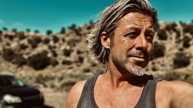 Bronz tenli, sakallı, atlet gömlekli, güneşli bir ortamda, arka planda siyah bir araba olan sarışın bir adam..