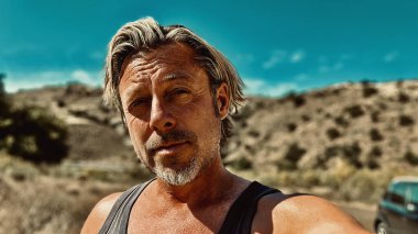 Bronz tenli, sakallı, atlet gömlekli, güneşli bir ortamda, arka planda siyah bir araba olan sarışın bir adam..