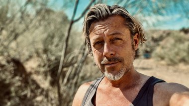 Bronz tenli, sakallı, atlet gömlekli, güneşli bir ortamda sarışın bir adam..