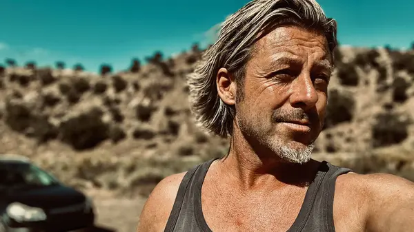 Bronz tenli, sakallı, atlet gömlekli, güneşli bir ortamda, arka planda siyah bir araba olan sarışın bir adam..