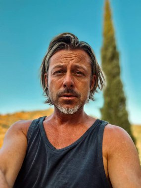 Bronz tenli, yarı sarı saçlı, sakallı, atlet tişörtlü, güneşli kırsalda tepeli, açık mavi gökyüzü olan bir adam..