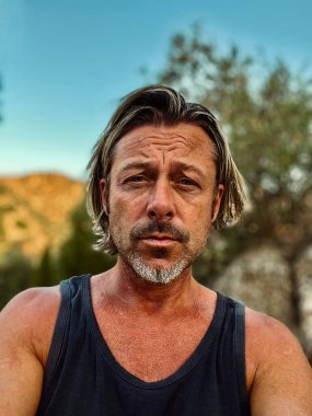Bronz tenli, yarı sarı saçlı, sakallı, atlet tişörtlü, güneşli kırsalda tepeli, açık mavi gökyüzü olan bir adam..