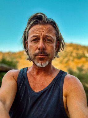 Bronz tenli, yarı sarı saçlı, sakallı, atlet tişörtlü, güneşli kırsalda tepeli, açık mavi gökyüzü olan bir adam..
