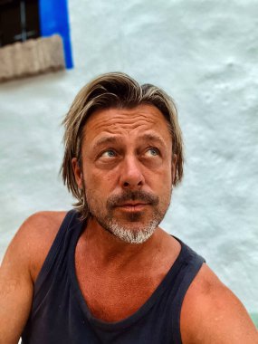 Koyu tenli, sarı saçlı, mavi gözlü, gri sakallı mavi atlet gömlekli, açık havada mavi pencereli beyaz duvarlı bir adam..