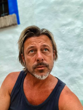 Koyu tenli, sarı saçlı, mavi gözlü, gri sakallı mavi atlet gömlekli, açık havada mavi pencereli beyaz duvarlı bir adam..