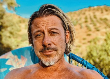 Gri sakallı, bronz tenli mutlu sarışın adam tepede ve güneşli kırsalda mavi bir sandalyede oturuyor..