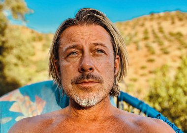 Gri sakallı, bronz tenli mutlu sarışın adam tepede ve güneşli kırsalda mavi bir sandalyede oturuyor..