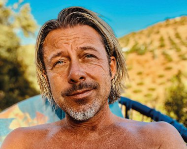 Gri sakallı, bronz tenli mutlu sarışın adam tepede ve güneşli kırsalda mavi bir sandalyede oturuyor..