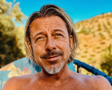 Gri sakallı, bronz tenli mutlu sarışın adam tepede ve güneşli kırsalda mavi bir sandalyede oturuyor..