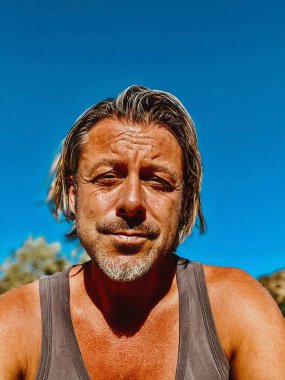Sarı saçlı, gri sakallı, mavi atletli mutlu bronzlaşmış adam açık havada, mavi gökyüzünün altında güneşli havanın tadını çıkar..