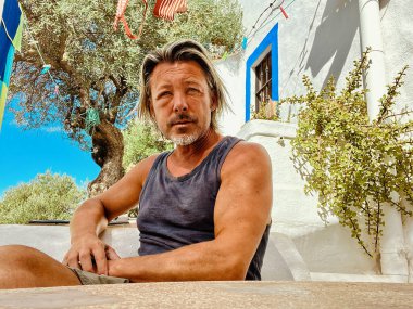 Bronzlaşmış sarışın bir adamın şişmiş yüzü gri sakallı, beyaz bir İspanyol evinin önünde oturuyor. Eşek arısı soktu..