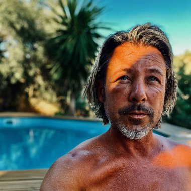 Bronzlaşmış, esmer, sakallı, sakalsız, yüzme havuzunun yanında oturan sarışın bir adamın şişmiş yüzü. Eşek arısı soktu..