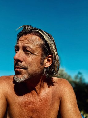 Bronz tenli, yarı sarı saçlı, gri sakallı, açık mavi gökyüzünün altında yaz güneşinde üstsüz bir adam..