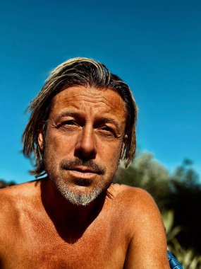 Bronz tenli, yarı sarı saçlı, gri sakallı, açık mavi gökyüzünün altında yaz güneşinde üstsüz bir adam..