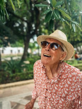 Senior man in red white shirt, straw hat and sunglasses walks outside in sunny city park of Ronda, Andalucia, Spain.