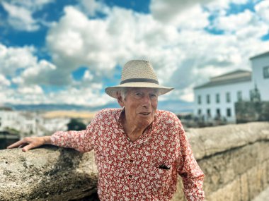 Senior man in red white shirt and straw hat stands outside on a bridge in Ronda, Andalucia, Spain.