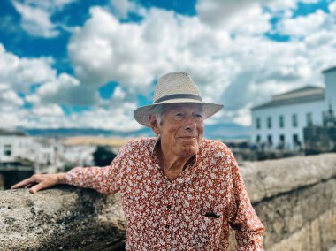 Senior man in red white shirt and straw hat stands outside on a bridge in Ronda, Andalucia, Spain.
