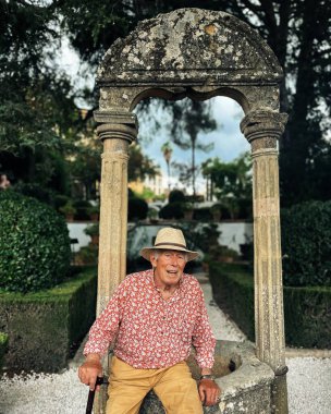 Senior man in red white shirt, straw hat and brown shorts sits on edge of water well in the garden of Casa del Rey Moro in Ronda, Andalucia, Spain.