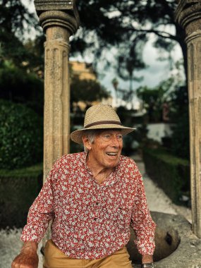 Senior man in red white shirt, straw hat and brown shorts sits on edge of water well in the garden of Casa del Rey Moro in Ronda, Andalucia, Spain.