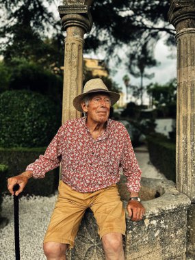 Senior man in red white shirt, straw hat and brown shorts sits on edge of water well in the garden of Casa del Rey Moro in Ronda, Andalucia, Spain.