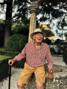 Senior man in red white shirt, straw hat and brown shorts sits on edge of water well in the garden of Casa del Rey Moro in Ronda, Andalucia, Spain.