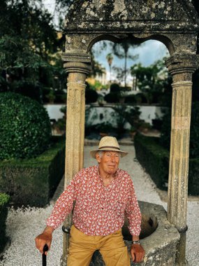 Senior man in red white shirt, straw hat and brown shorts sits on edge of water well in the garden of Casa del Rey Moro in Ronda, Andalucia, Spain.
