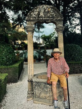 Senior man in red white shirt, straw hat and brown shorts sits on edge of water well in the garden of Casa del Rey Moro in Ronda, Andalucia, Spain.
