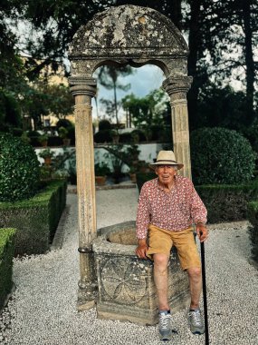 Senior man in red white shirt, straw hat and brown shorts sits on edge of water well in the garden of Casa del Rey Moro in Ronda, Andalucia, Spain.