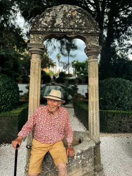 Senior man in red white shirt, straw hat and brown shorts sits on edge of water well in the garden of Casa del Rey Moro in Ronda, Andalucia, Spain.